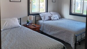 Une chambre avec deux lits, une table de chevet avec une lampe, un tableau encadré et une fenêtre avec des stores.