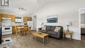 A modern kitchen with wooden cabinets, a dining area with a table and chairs, a gray sofa with patterned cushions, and a painting on the wall.
