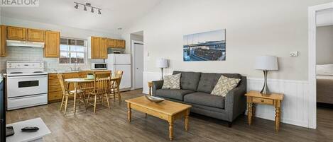 A modern kitchen with wooden cabinets, a dining area with a table and chairs, a gray sofa with patterned cushions, and a painting on the wall.