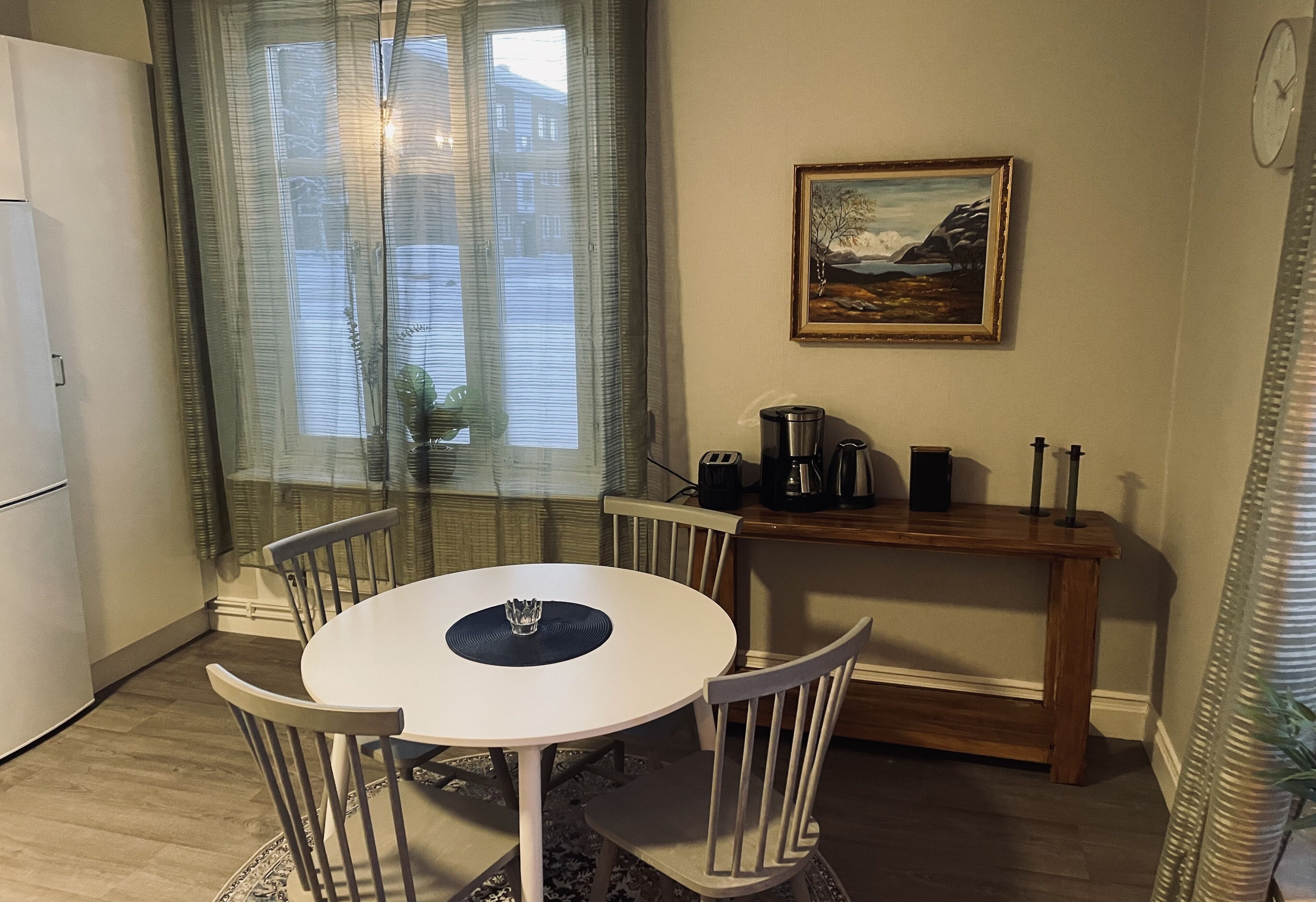 Una zona de menjador petita amb una taula blanca i quatre cadires, una taula auxiliar de fusta amb una cafetera, un quadre a la paret i una finestra amb cortines transparents.