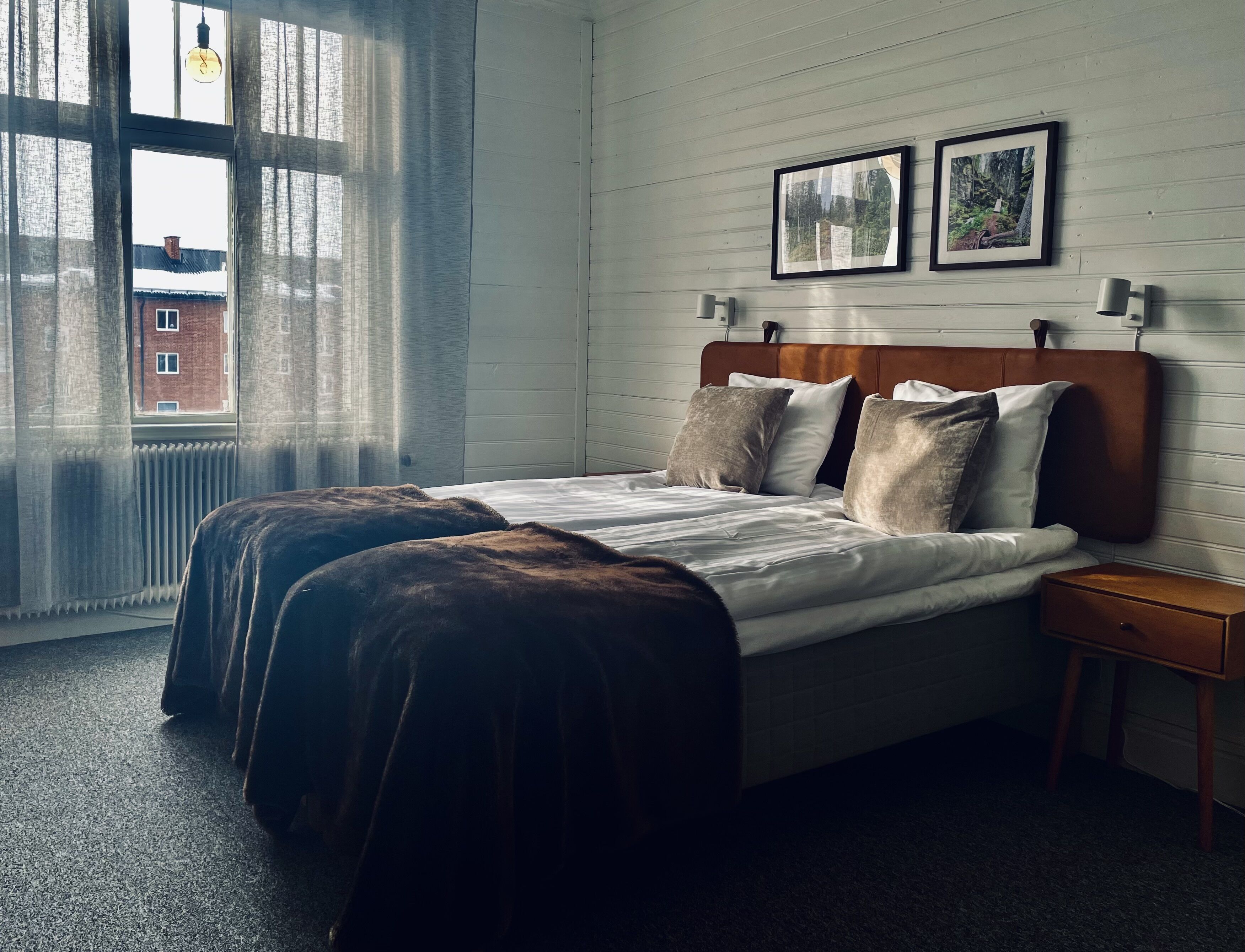 A hotel room with a large window, a bed with a wooden headboard, two framed pictures on the wall, and a bedside table.