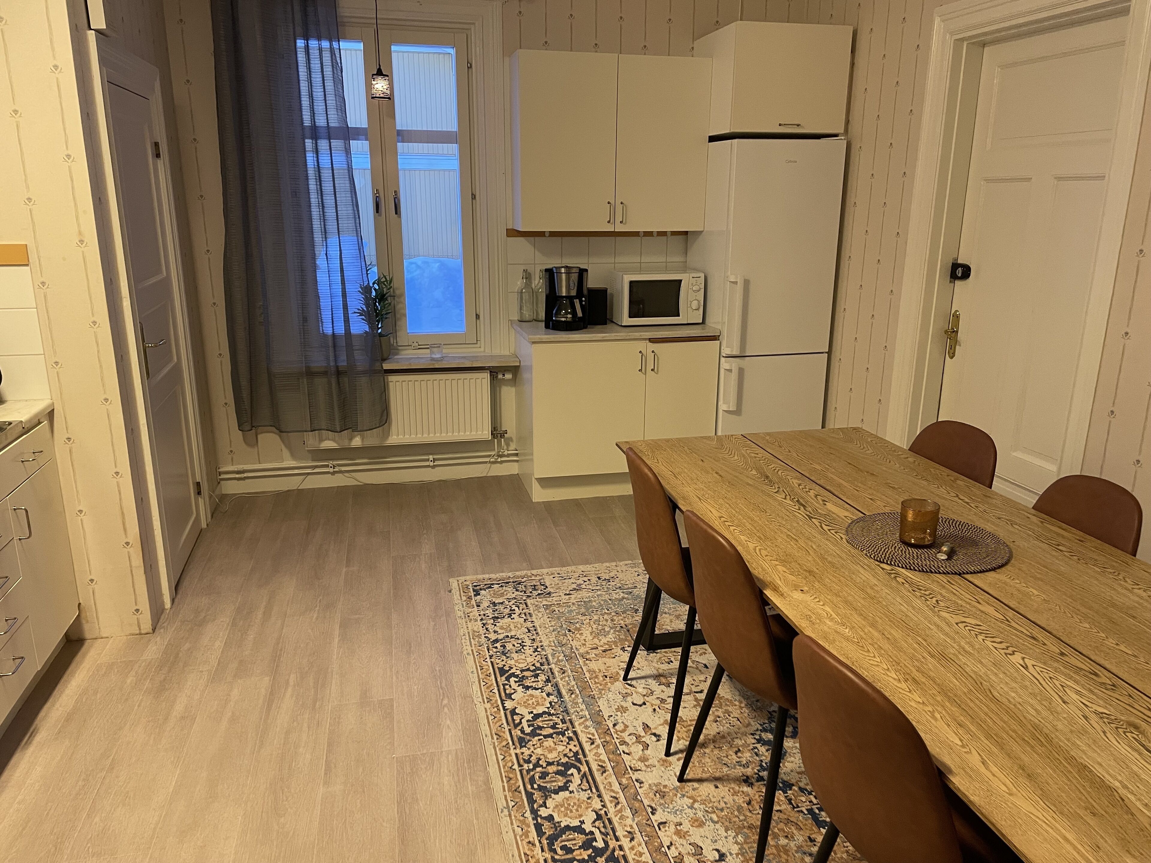 Uma cozinha compacta com armários brancos, uma mesa de jantar de madeira e um tapete estampado.