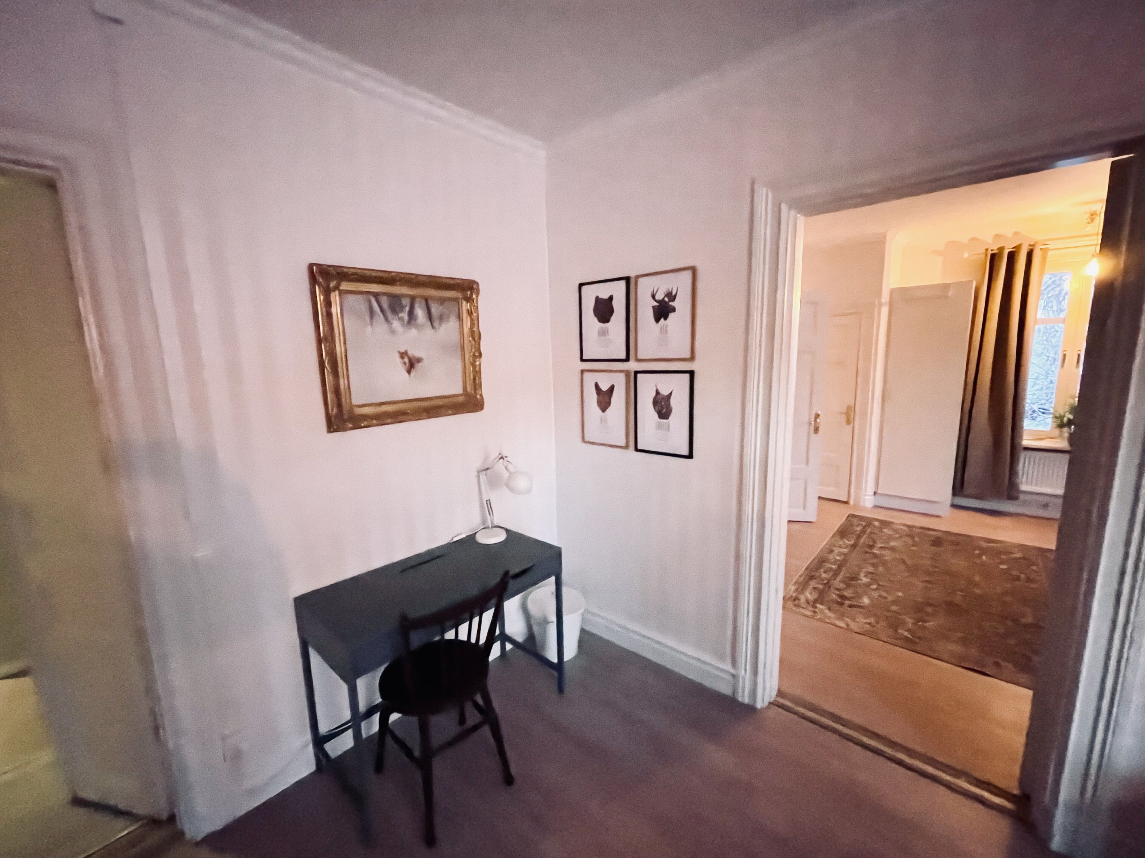 A room with a desk, chair, framed pictures on the wall, and a doorway leading to another room.