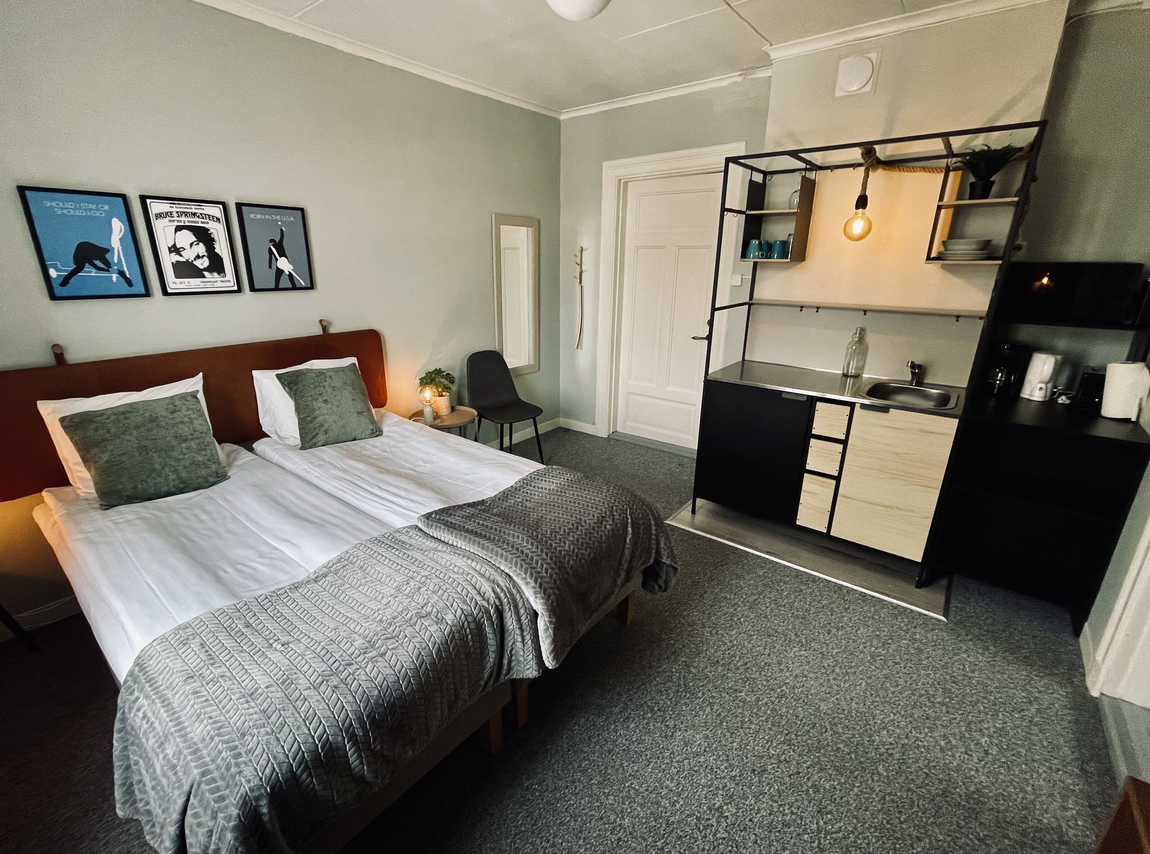 Une chambre d’hôtel comprenant un lit, un bureau, une chaise, une petite table, un lavabo et une étagère avec une plante.