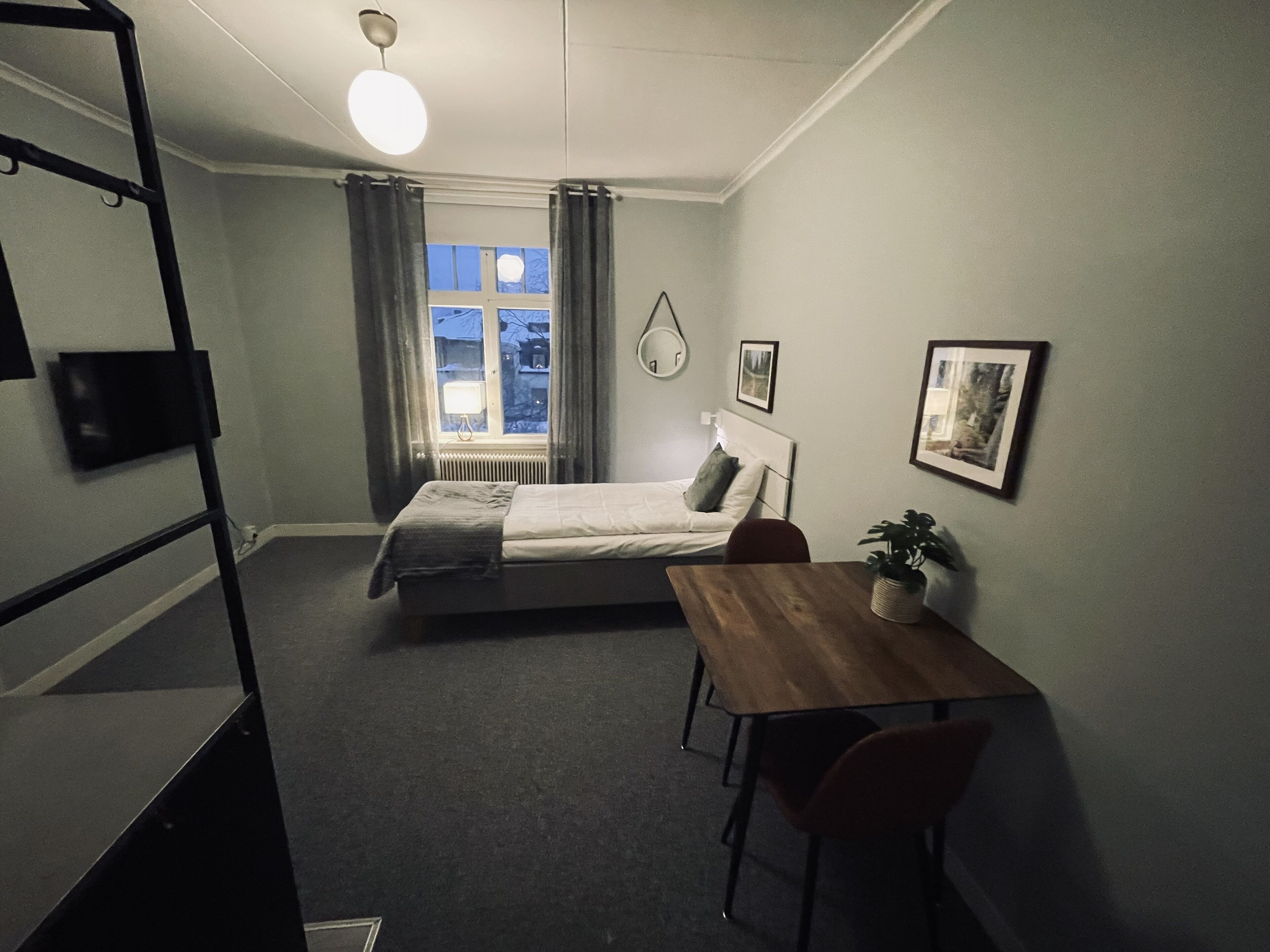 A small, well-lit bedroom with a single bed, a desk, and a chair. There is a window with curtains, a radiator, and a wall-mounted TV.