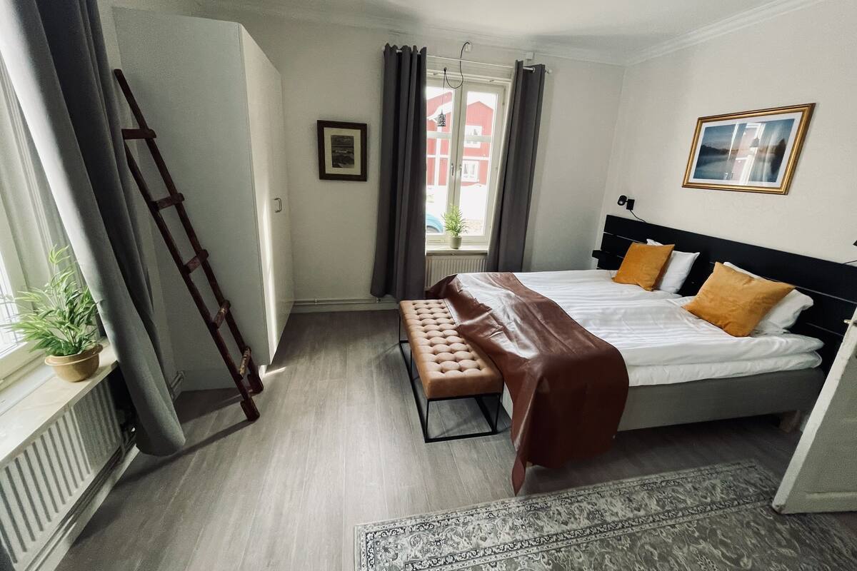 Un dormitorio con cama, un banco, un radiador, una ventana con cortinas y un cuadro en la pared.
