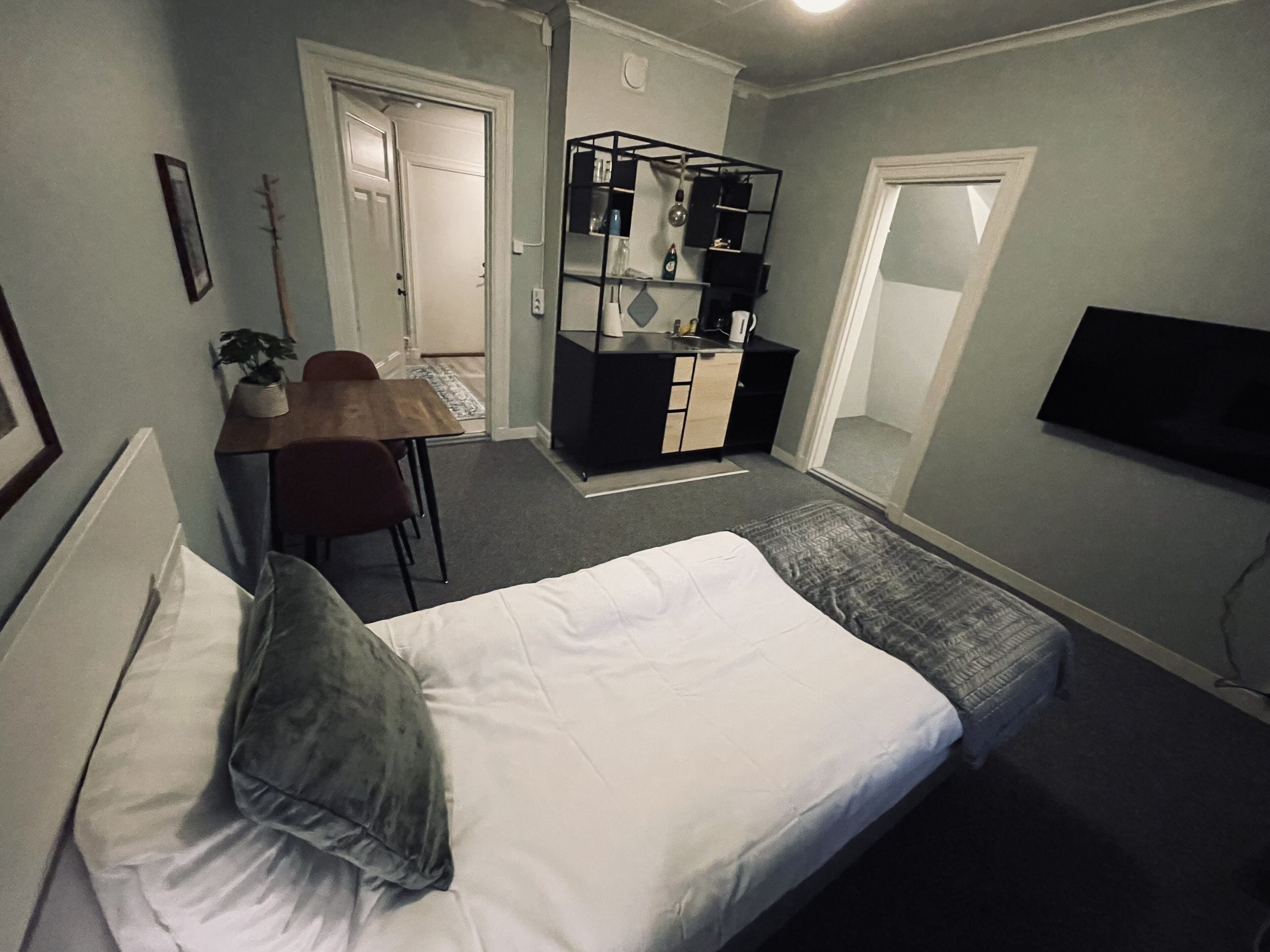 A small, compact room with a bed, a desk, and a chair. There is a TV mounted on the wall and a door leading to another room.
