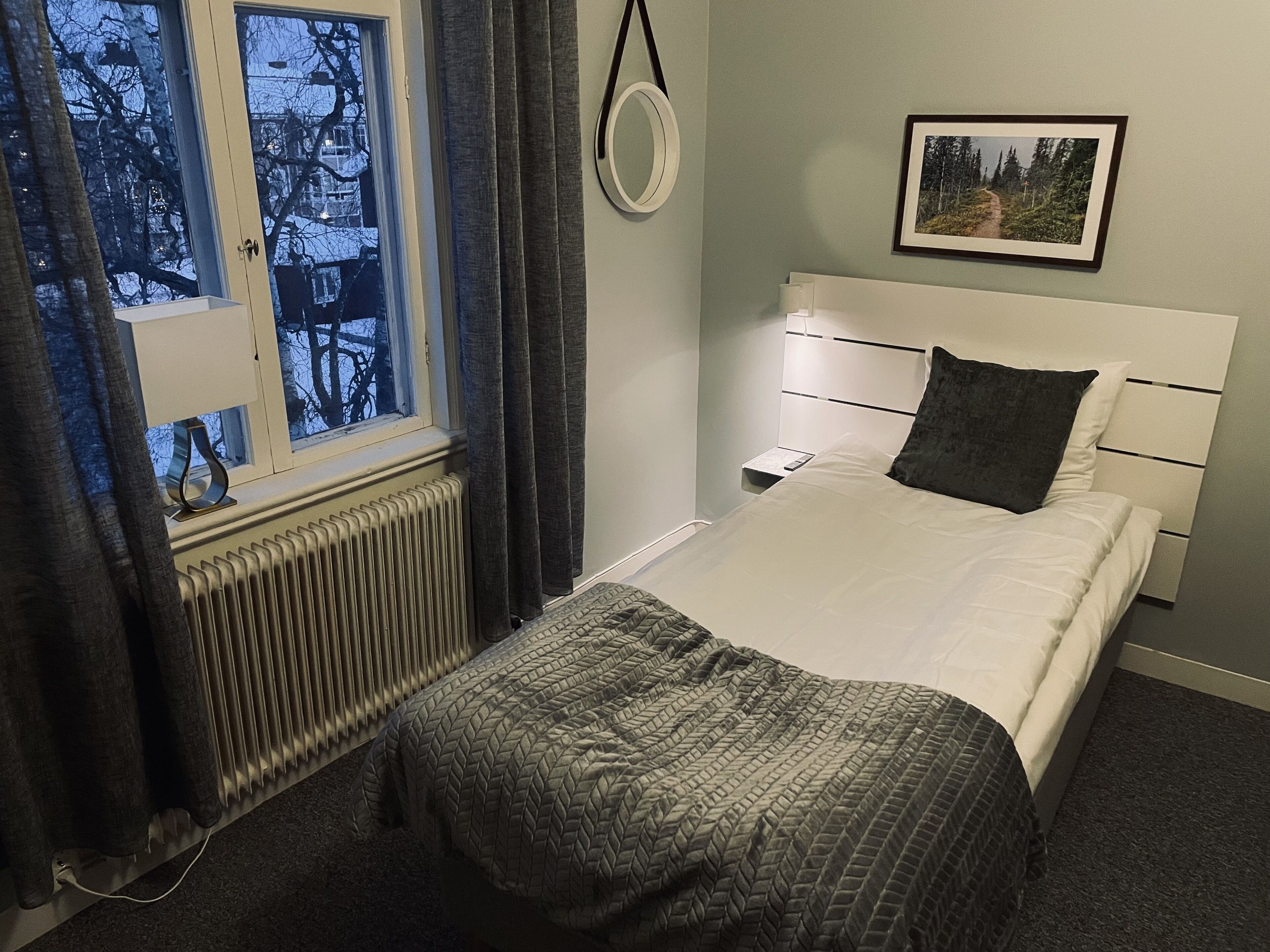 Une chambre à coucher comprenant un lit, une table de chevet, une fenêtre avec des rideaux, un radiateur et un tableau encadré accroché au mur.