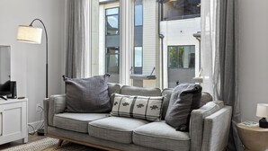 Una sala de estar moderna con un sofá gris, una lámpara de pie y vistas a un edificio a través de amplios ventanales.