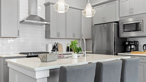 Cocina moderna con gabinetes blancos, electrodomésticos de acero inoxidable, una isla central con taburetes y luminarias colgantes.
