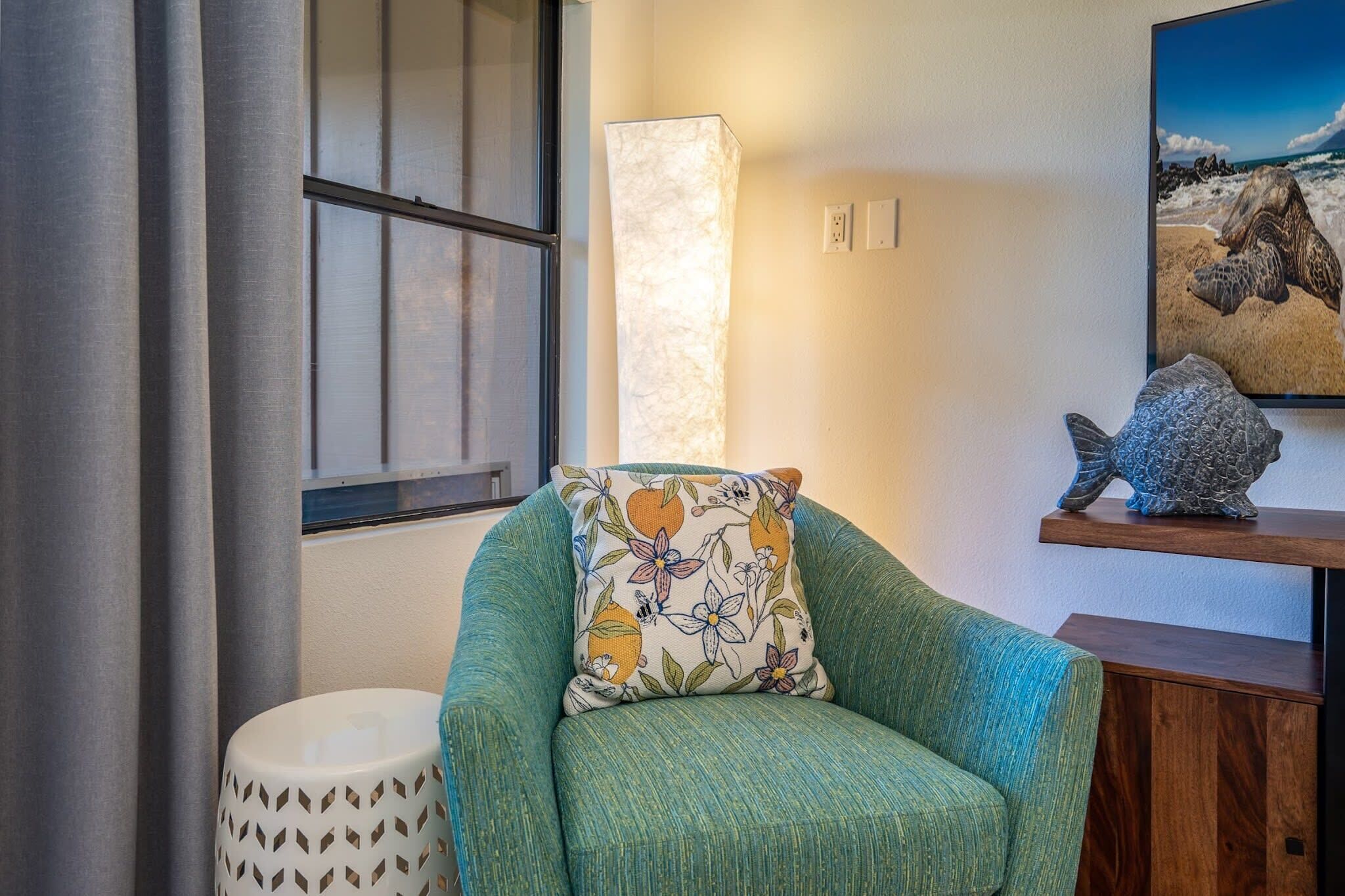 A teal armchair with a floral pillow, a white perforated stool, a tall white lamp, and a wooden shelf with a turtle sculpture.