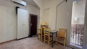 Una habitación pequeña con suelo de baldosas, una puerta de madera, una mesa con sillas, una lámpara y una ventana con persianas.