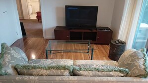 Un salon comprenant un canapé, une table basse en verre, un téléviseur à écran plat posé sur un meuble en bois, et une fenêtre avec des rideaux.