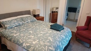 Une chambre à coucher avec un lit, une table de chevet, un fauteuil rouge et une salle de bain visible à travers une porte ouverte.