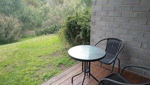Une terrasse avec une table ronde et deux chaises, un mur en briques et une zone herbeuse agr émentée d’arbustes.