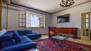 A living room with a sectional sofa, a patterned rug, a glass coffee table, a wooden cabinet, and a large window with open shutters. - Nice home in Terrasini (Terrasini)