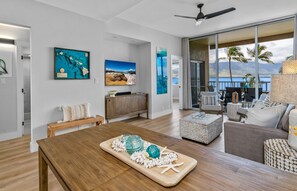 A modern living room with a wooden dining table, a sofa, and a large window overlooking a scenic view. - Upscale Oceanfront Penthouse Vistas (Kihei)