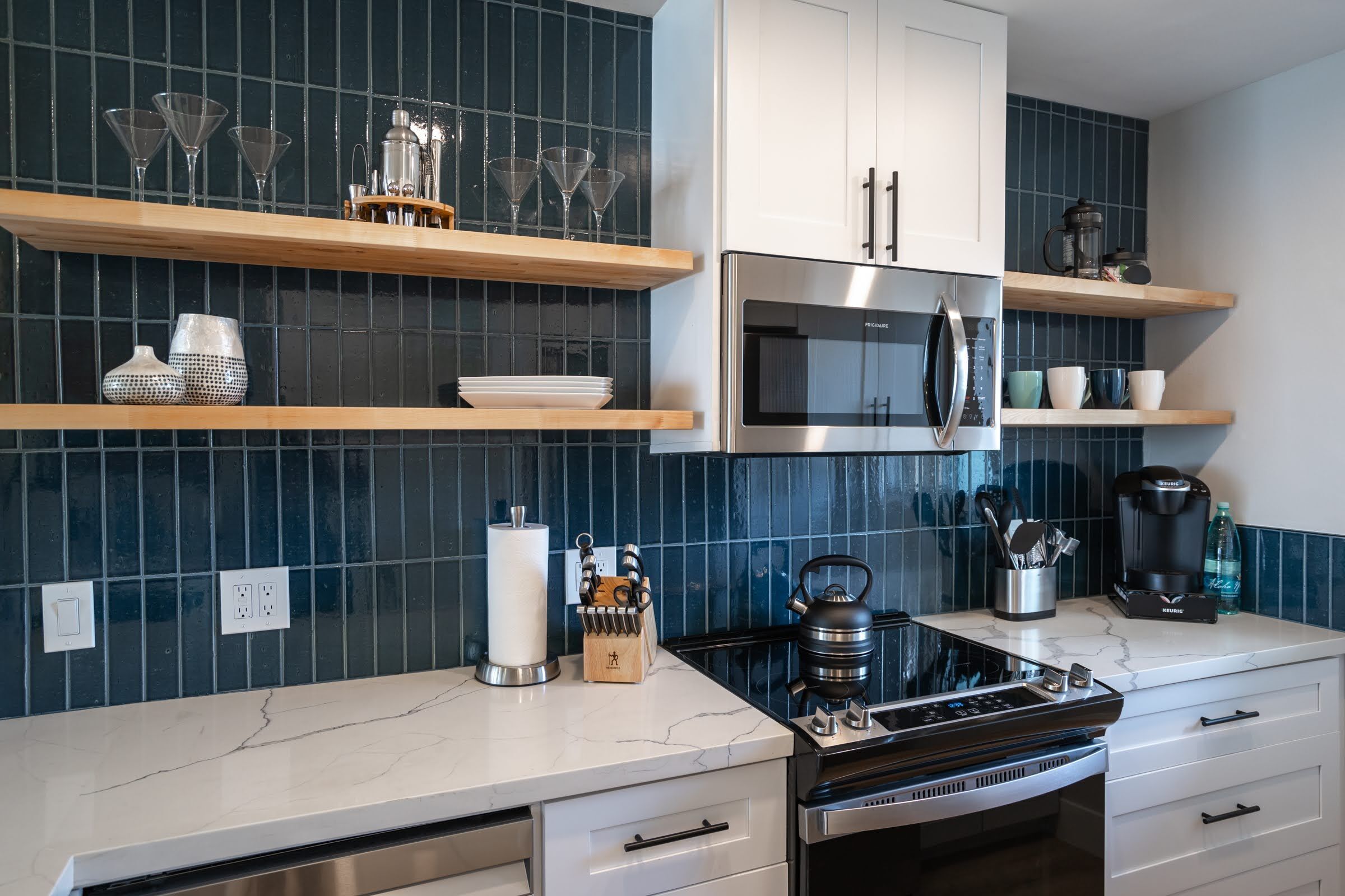 Una cocina moderna con refrigerador de acero inoxidable, microondas y estufa. Hay estantes de madera con cristalería y utensilios de cocina.