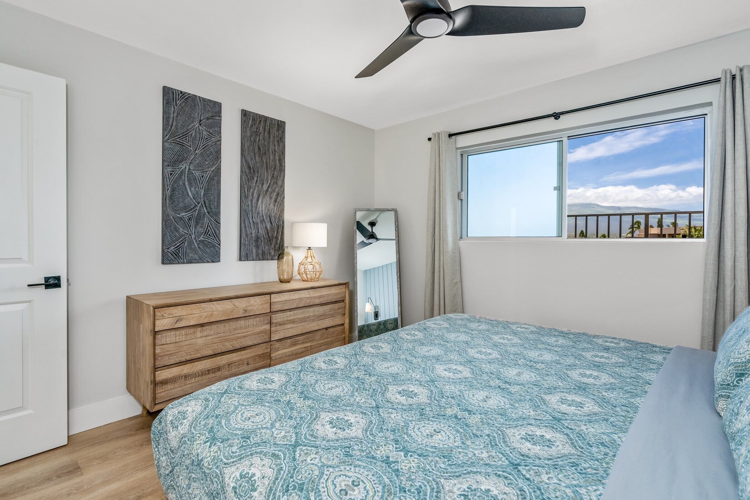 A bedroom with a bed, a ceiling fan, a window with a view, a dresser, and two wall art pieces.
