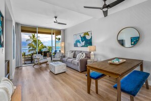 A spacious living area with a wooden floor, a grey sofa, a wooden dining table with blue cushions, and a view of the ocean through large windows. - Upscale Oceanfront Penthouse Vistas (Kihei)