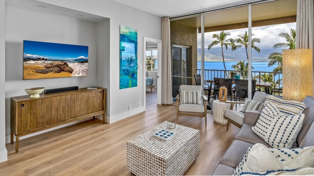 A modern living room with a large TV, wooden console, and a view of the ocean through large sliding doors. - Upscale Oceanfront Penthouse Vistas (Kihei)