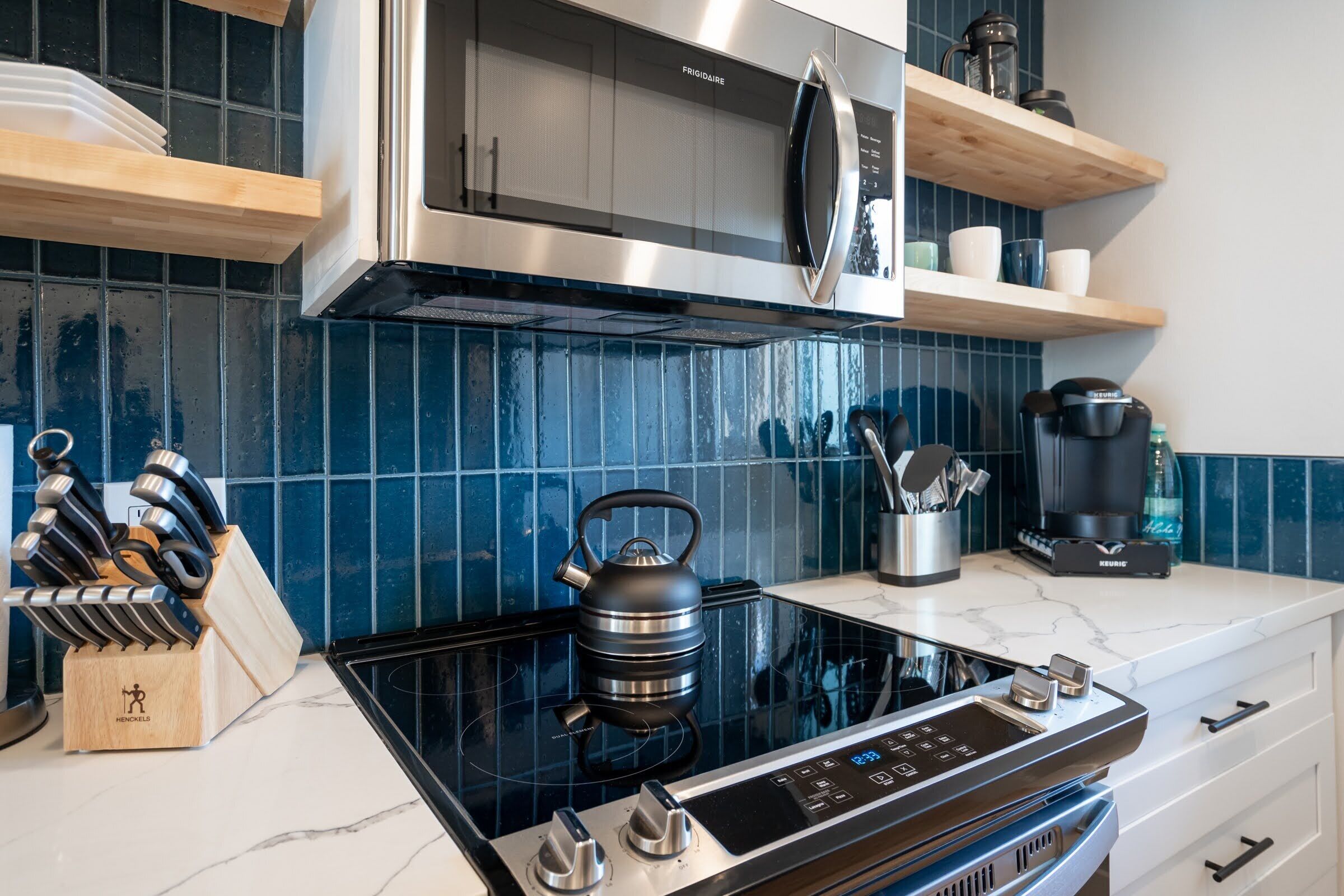 A modern kitchen with a black induction cooktop, stainless steel appliances, and blue-tiled backsplash.