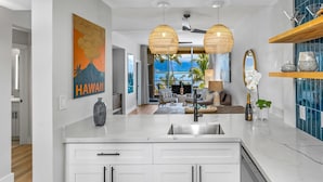 A modern kitchen with white cabinetry, a large island, and a view of a balcony with palm trees. - Upscale Oceanfront Penthouse Vistas (Kihei)