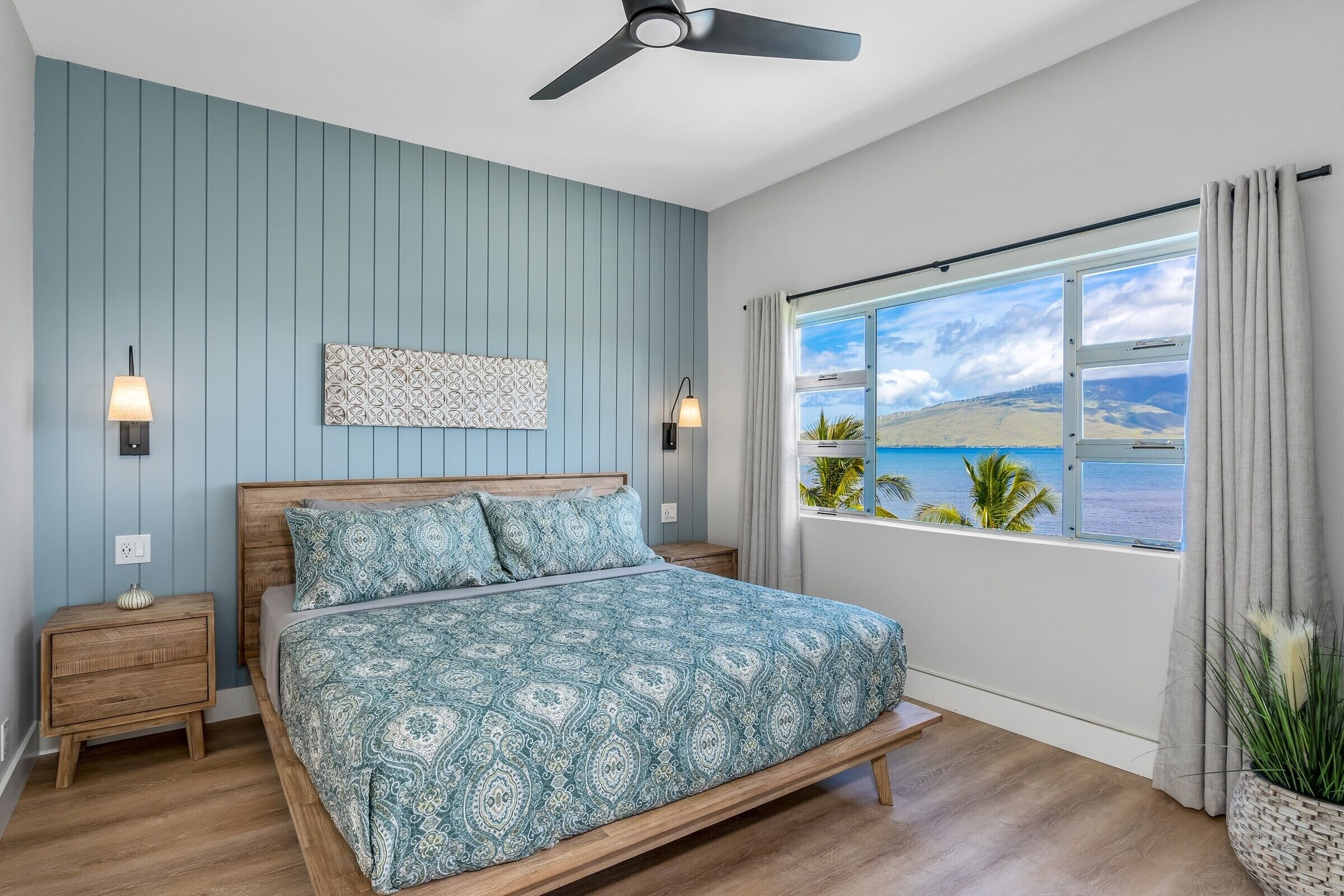 A bedroom with a bed, wooden nightstands, a ceiling fan, a window with a view of the sea, and a decorative wall piece.