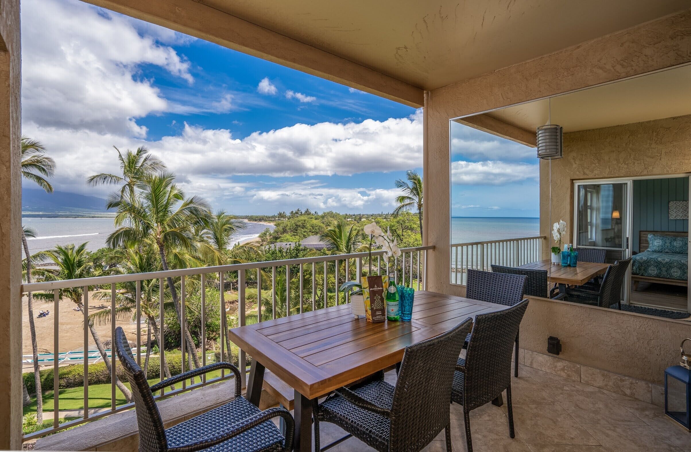 Ein Balkon mit Holztisch und Korbstühlen mit Blick auf einen Strand und ein Meer mit Palmen.