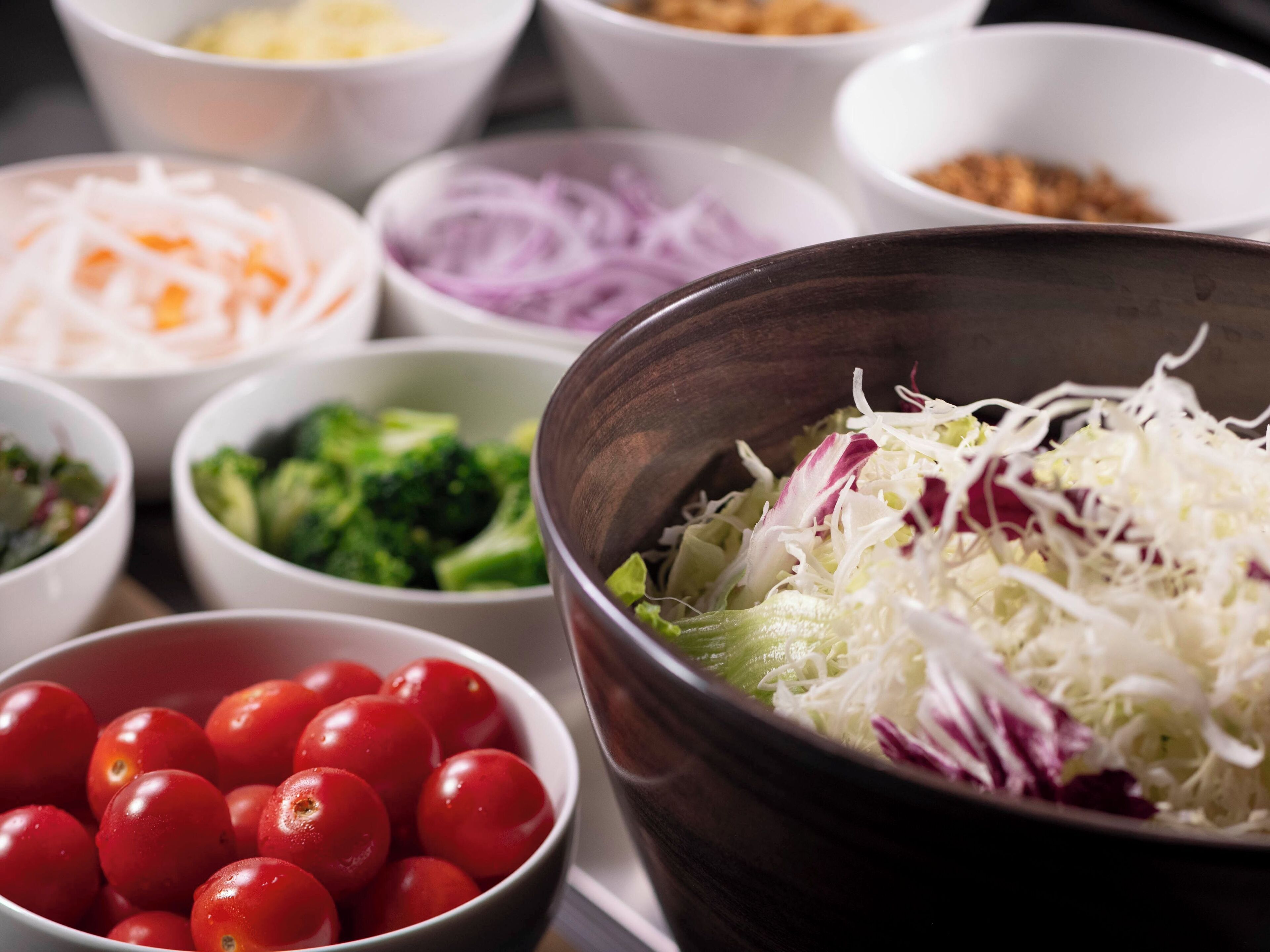 Verschillende soorten eten in kommen, zoals geraspte kool, rode uien, broccoli, kerstomaatjes en andere ingrediënten.