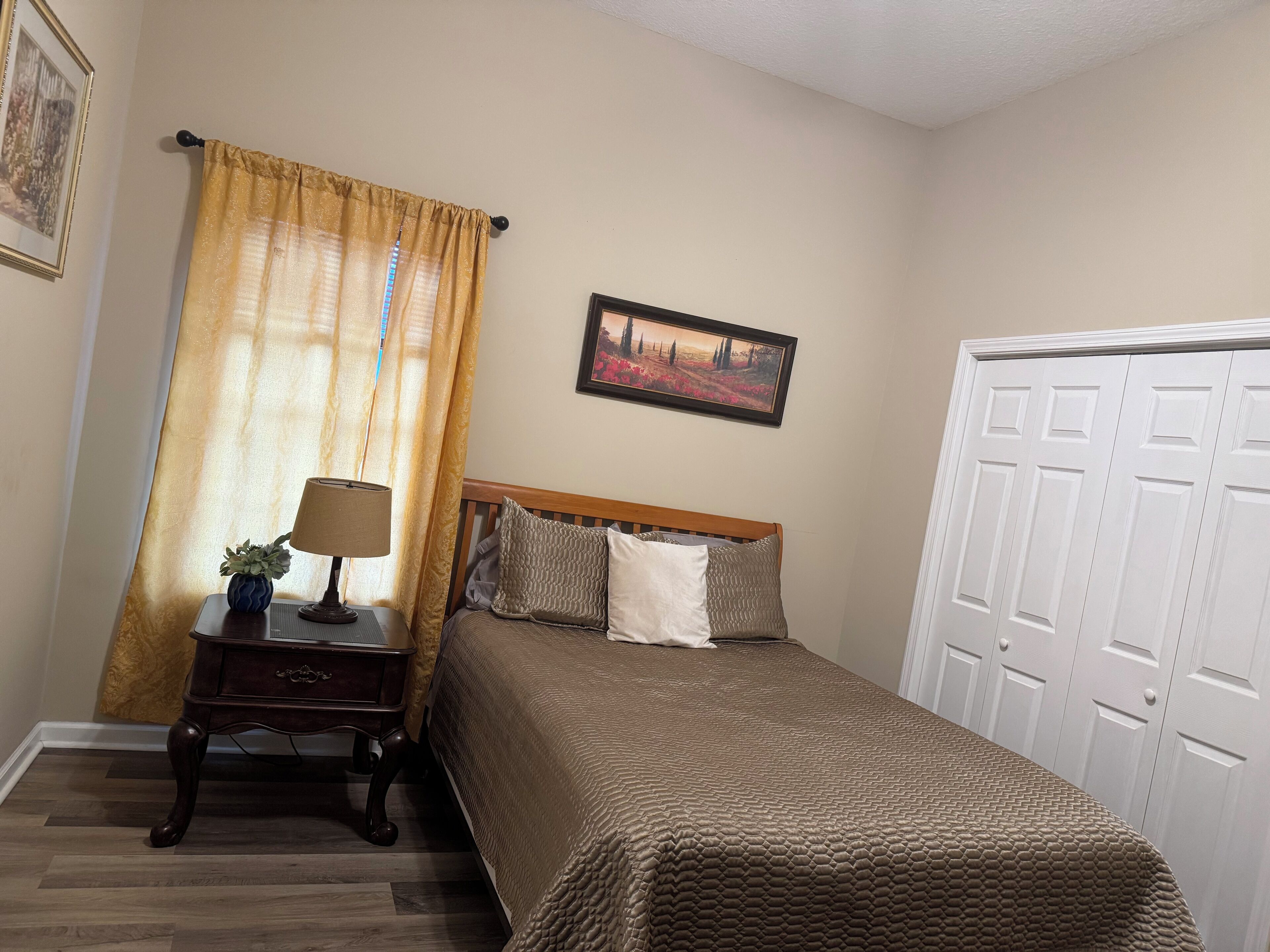 A bedroom with a bed, a nightstand, a lamp, a window with curtains, a framed picture on the wall, and a door.