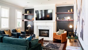 A living room with a fireplace, blue and green sofas, a television, and shelves with decorative items. - Luxury Design, Prime Location 2 Minutes from Midtown, Family Fun! (Memphis)