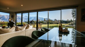 Un comedor moderno con una mesa y sillas de vidrio, ventanas amplias que ofrecen vistas a la montaña, y una zona de estar acogedora con un sofá y cojines.