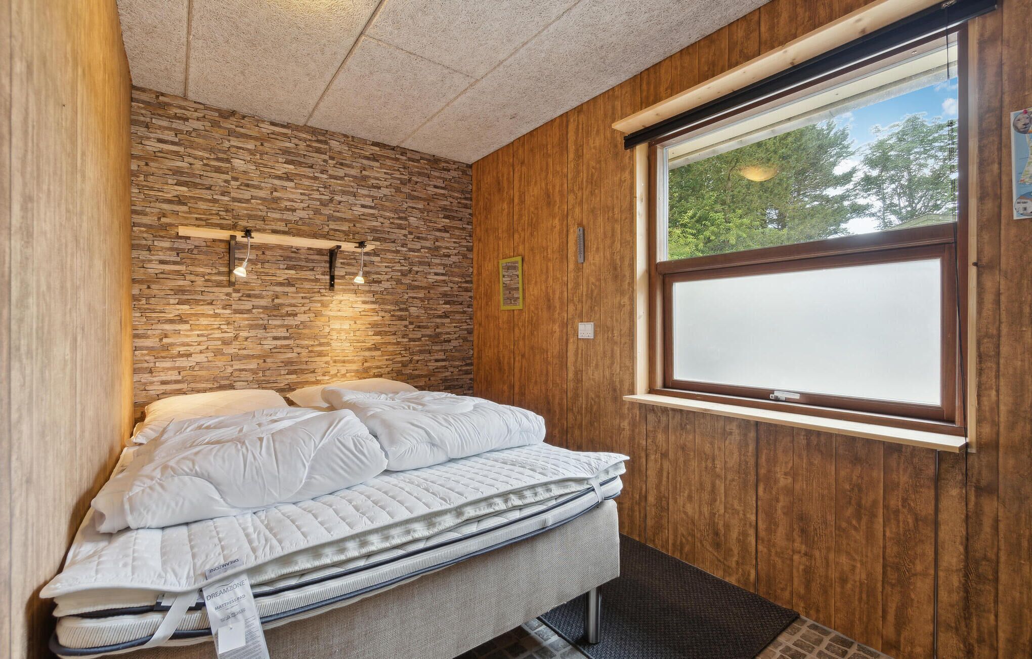 Quarto com parede de pedra, painéis de madeira, uma janela grande e cama com roupa de cama branca.