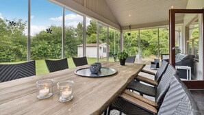 A covered patio with a wooden table, wicker chairs, and a view of a lush garden.