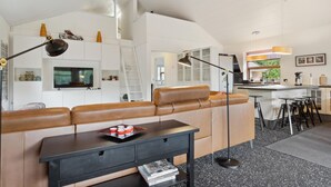A modern living room with a leather sofa, a black side table, and a TV. There is a kitchen area with a bar and stools.