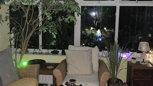 Ein gemütlicher Innenbereich mit einem Korbssessel, einem gepolsterten Sessel und einem kleinen Tisch mit Lampe. Ein großes Fenster gibt den Blick auf einen Garten mit Baum und dekorativer Beleuchtung frei.