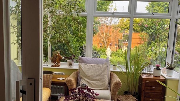 Ein gemütliches Zimmer mit einem Korbstuhl, einem kleinen Tisch mit Blumenvase und Blick auf einen Garten durch große Fenster.