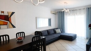 A modern living room with a dark sofa, a dining table with chairs, a contemporary light fixture, and a large window with curtains. - T3  Familiar Albufeira, wifi, a/c, praia a 500m (Albufeira)