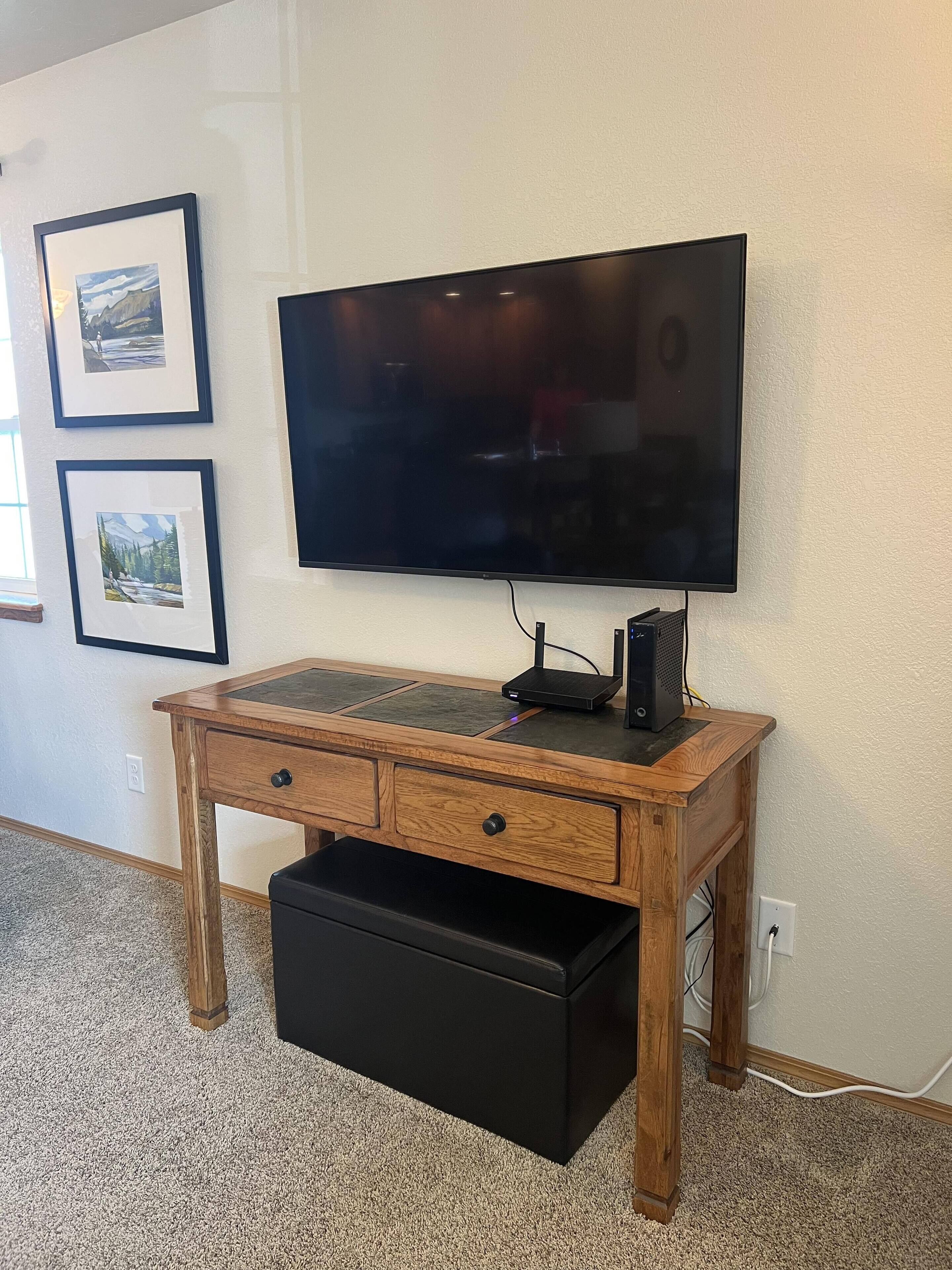 A wooden desk with a flat-screen TV mounted on the wall, a black drawer unit, and two framed pictures on the wall.