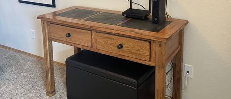 A wooden desk with a flat-screen TV mounted on the wall, a black drawer unit, and two framed pictures on the wall.