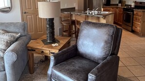En stue med sofa, læderlænestol, lampe og spiseområde med træskabe og et køleskab.