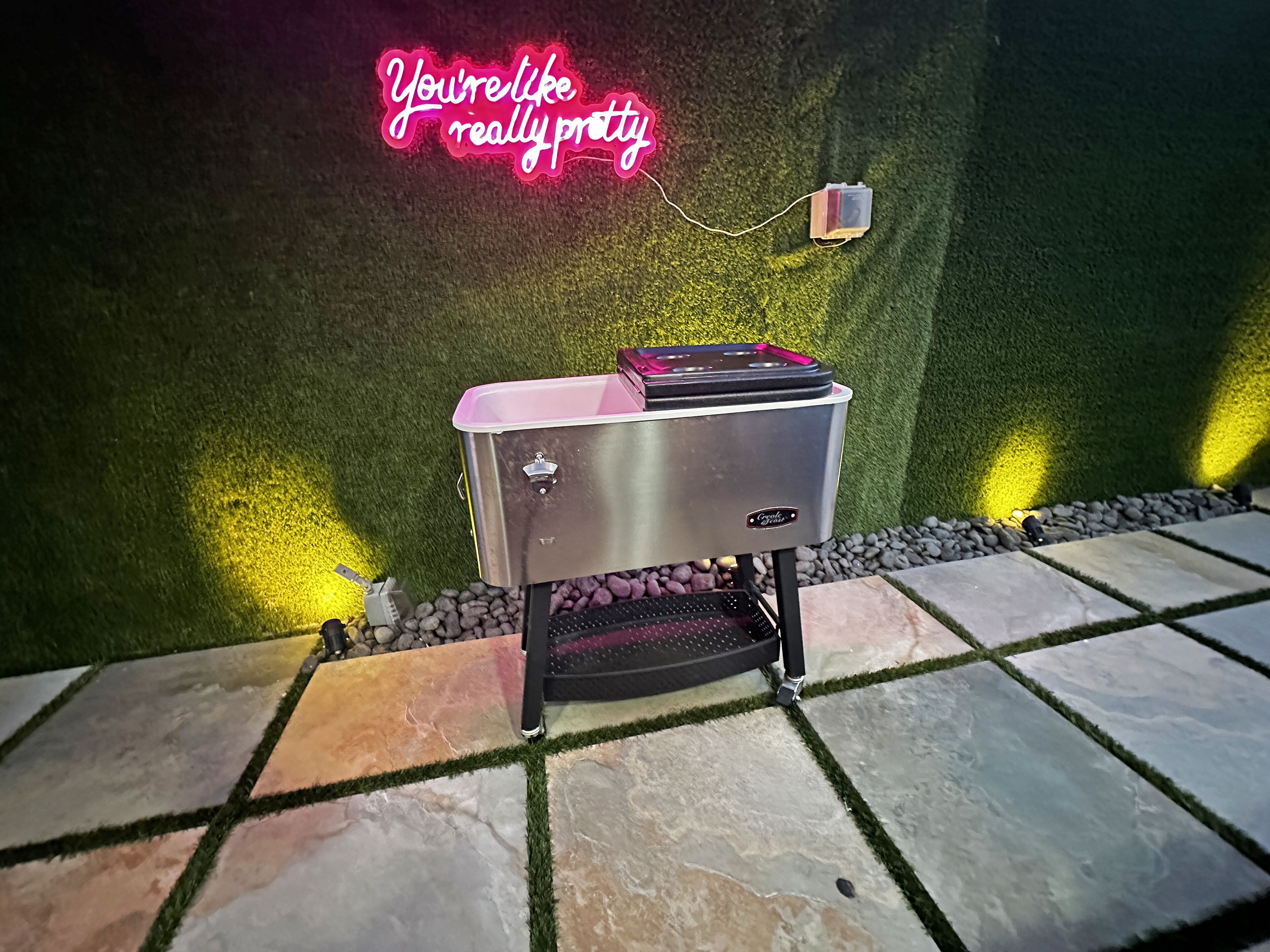 Un barbecue portable doté d’un couvercle rose et d’un corps en acier inoxydable, placé sur une terrasse carrelée en pierre, bordée d’une pelouse décorative.