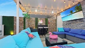 Ein Außenbereich mit blauen Sofas, einem rosa Flamingo-Stuhl und einem an der Wand befestigten Fernseher, der eine Strandansicht zeigt.