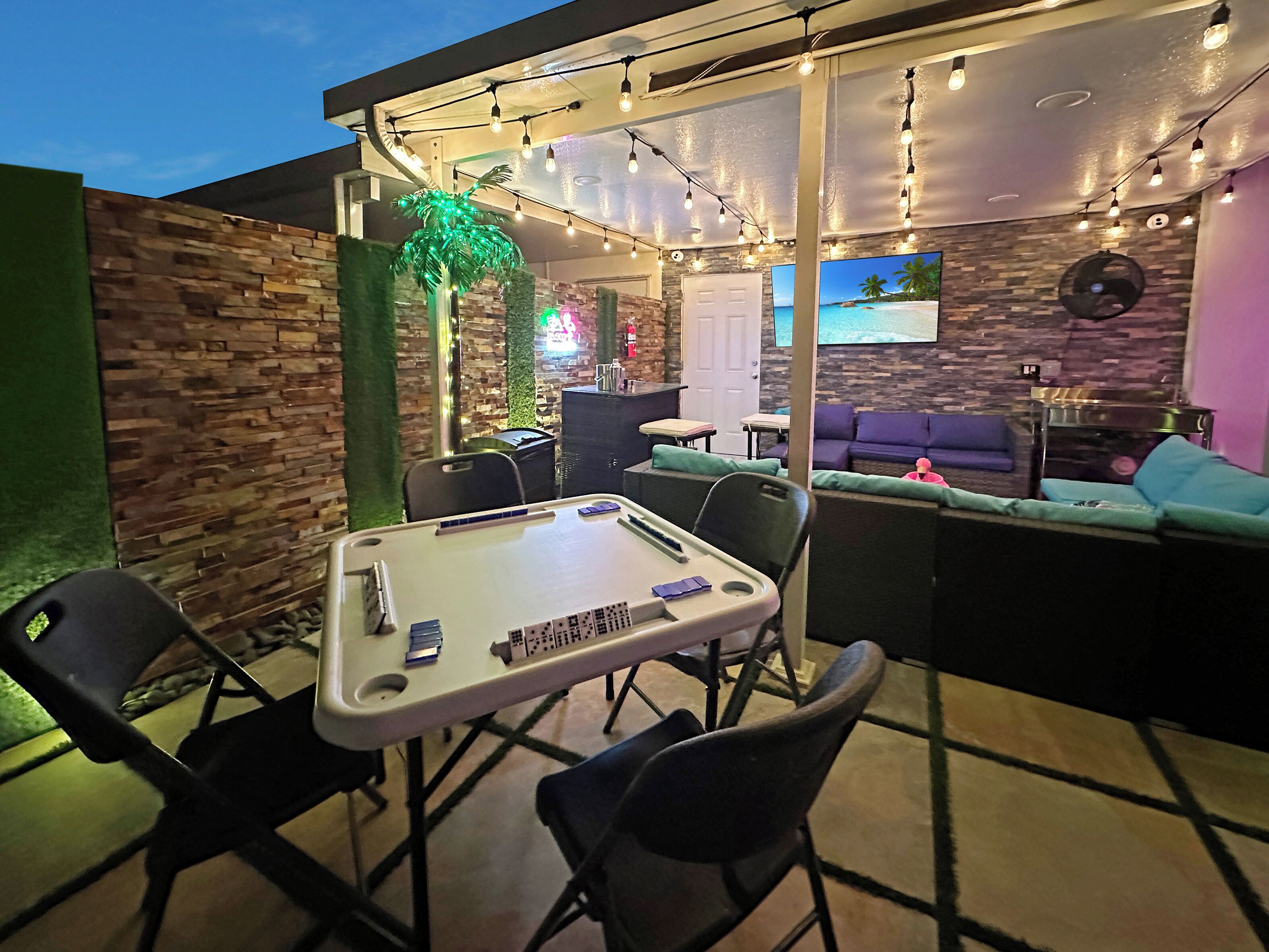 Un espace extérieur aménagé avec une table de billard, des chaises et un écran de télévision diffusant des images d’une plage.