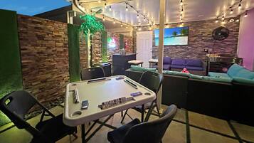 Un espace extérieur aménagé avec une table de billard, des chaises et un écran de télévision diffusant des images d’une plage.