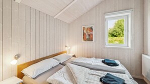 A bedroom with a wooden ceiling, a bed with white bedding, a nightstand, a wall-mounted lamp, a window with a view of greenery, and a painting on the wall. - Cozy home in Otterndorf with sauna (Otterndorf)