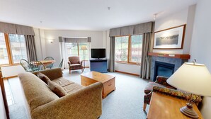 Een woonkamer met een bank, fauteuil, salontafel, televisie, open haard en grote ramen.