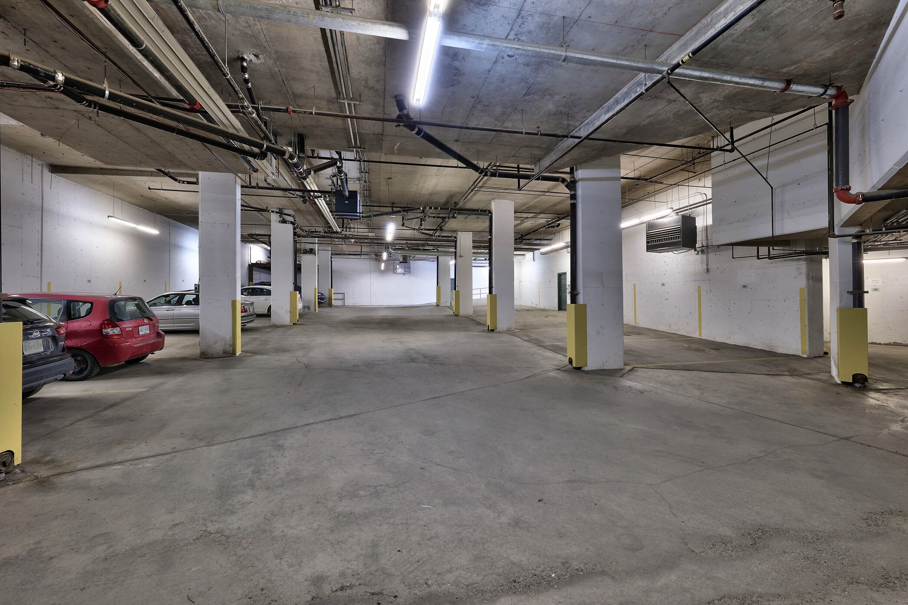 En parkeringskælder med betongulv og -søjler, lysrør og synlige rør og kanaler.