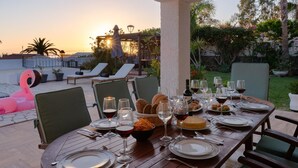 Een terras met eethoek en een zonsondergang, tafels gedekt met wijnglazen, borden en brood, en lounge fauteuils op de achtergrond.